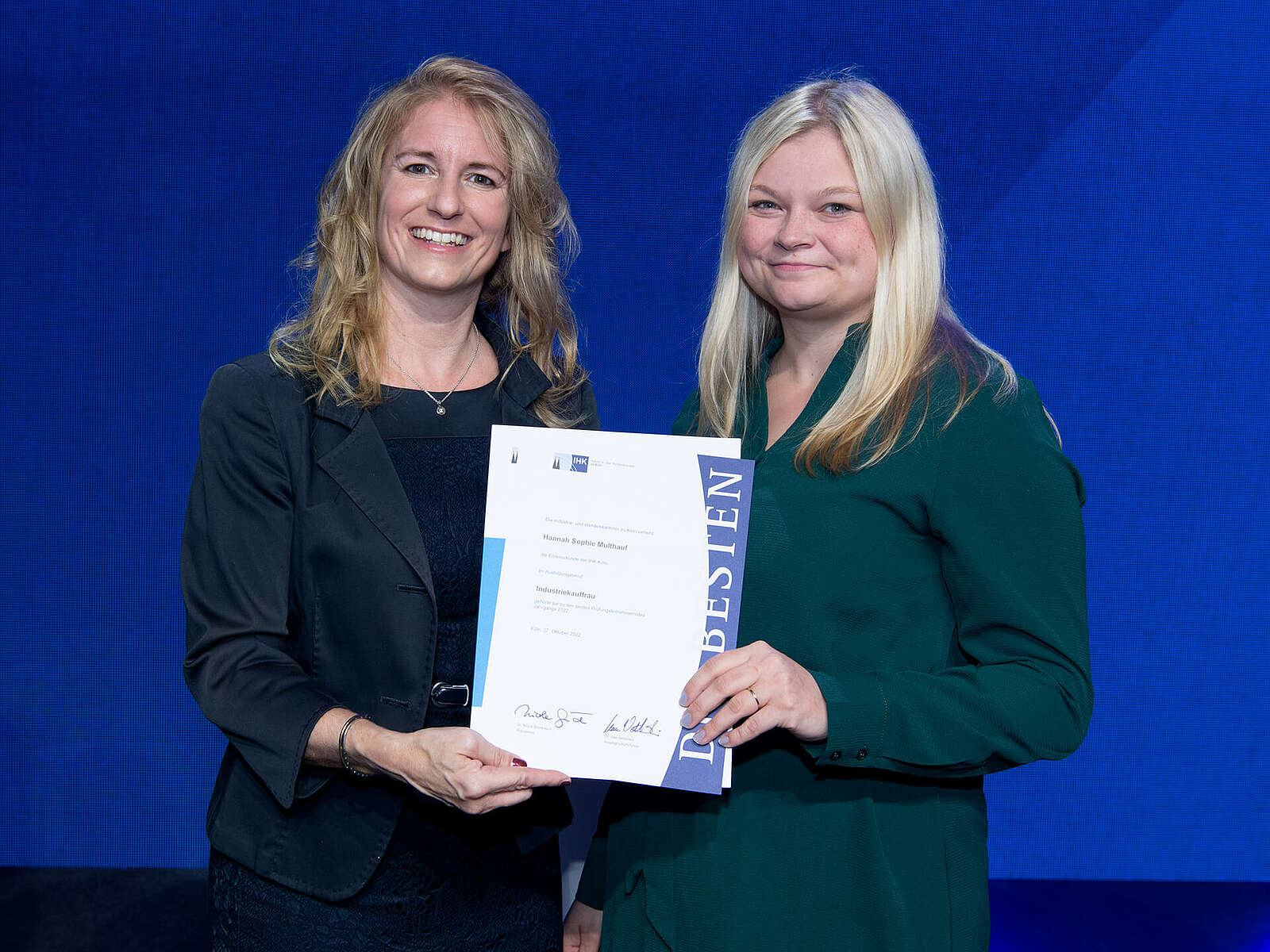 Two women smiling proudly at the camera while holding an IHK certificate for a successful vocational training.
