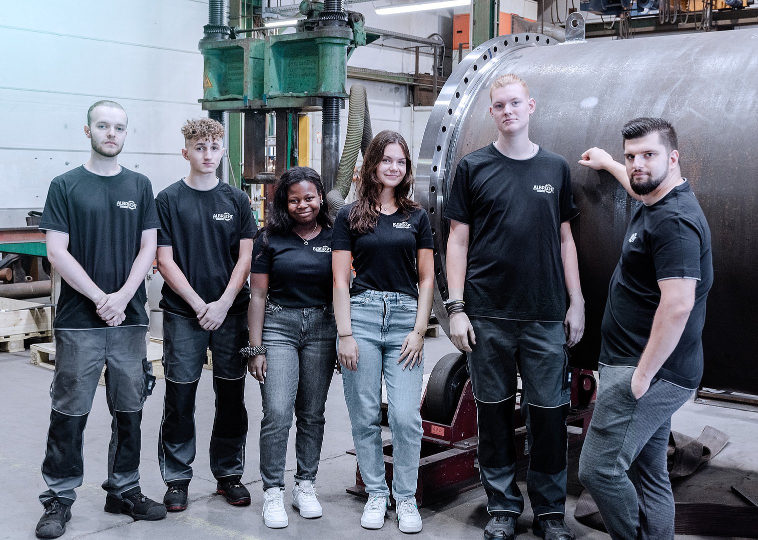 Six young workers in black t-shirts and work pants standing proudly inside an industrial factory with machinery and large metal parts.