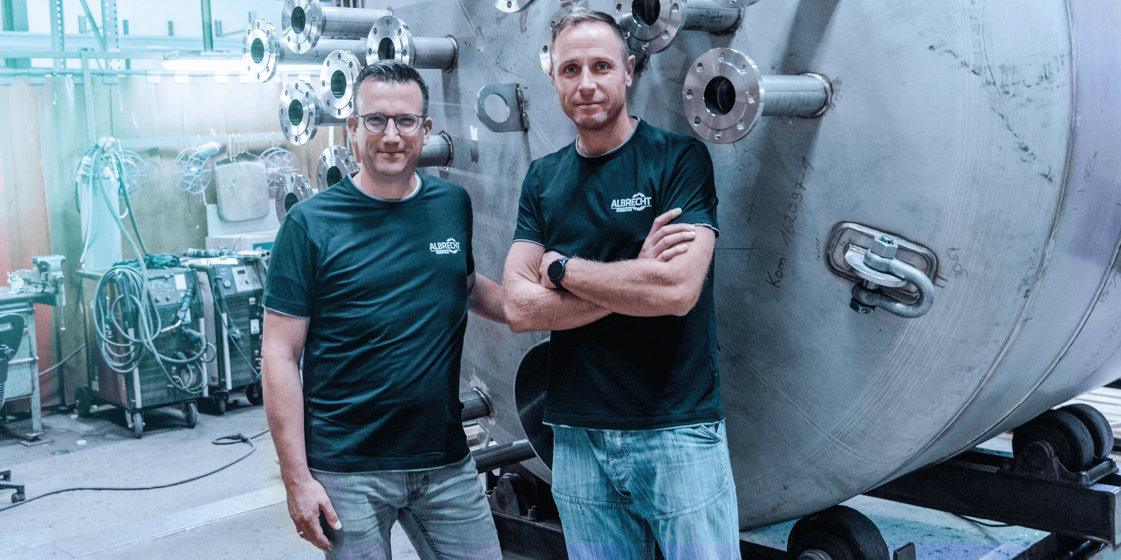 Two men in dark shirts stand in a workshop in front of a large stainless steel industrial tank.