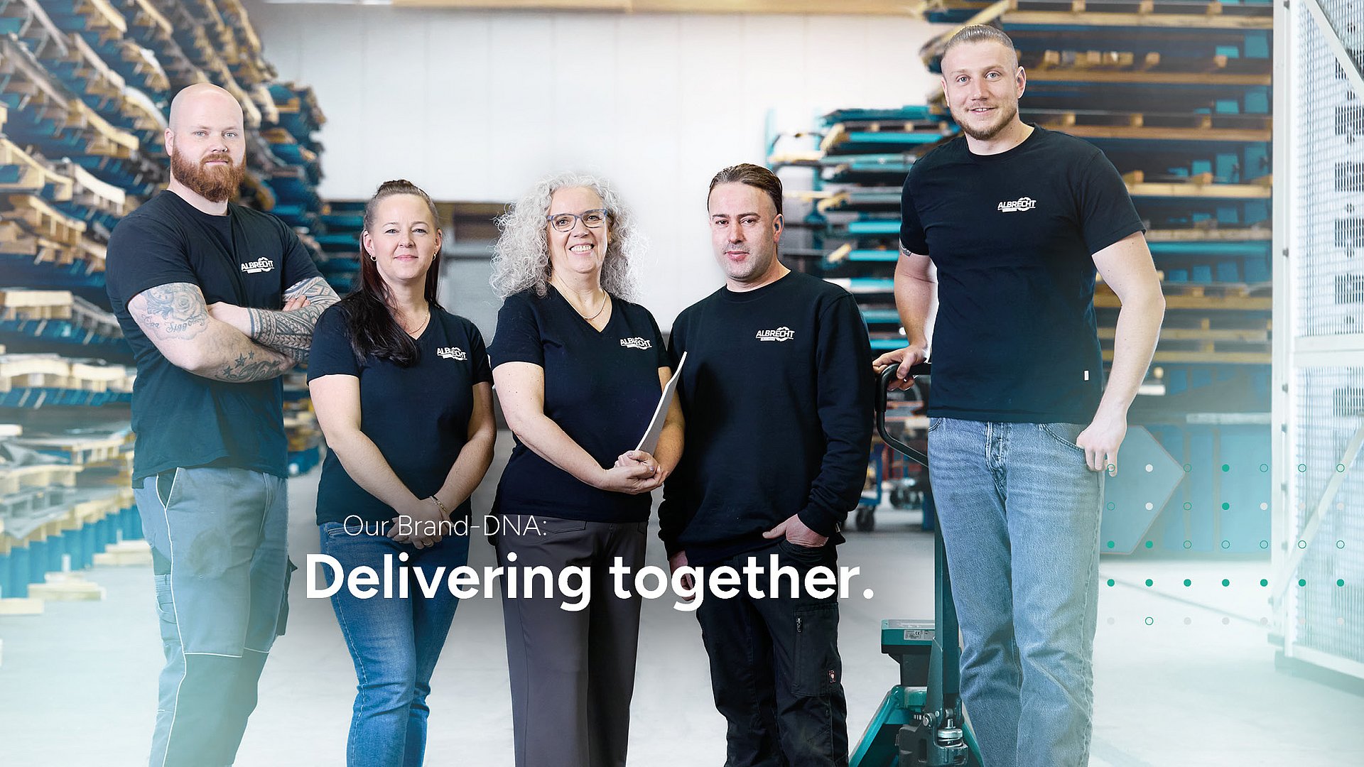 Albrecht Team in warehouse Five cheerful employees of Albrecht stand in a warehouse in front of metal shelves and smile at the camera.