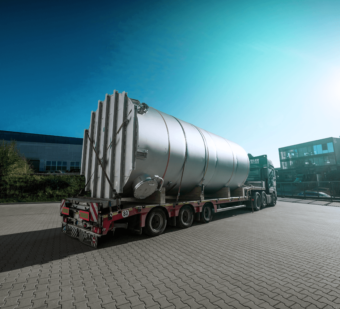 Ein schwerer Lkw transportiert einen riesigen Metallsilo auf einem Auflieger über einen gepflasterten Hof.