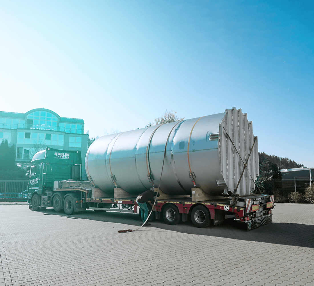 Ein Lkw transportiert ein großes zylindrisches Silo, das auf einem Tieflader mit Gurten gesichert wird.