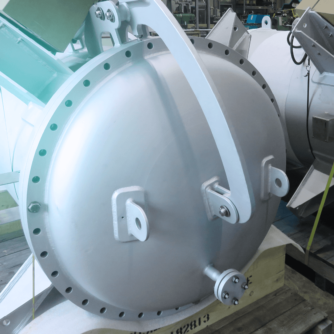 A large, bright metal industrial airlock with a flange and lifting lugs in a workshop.