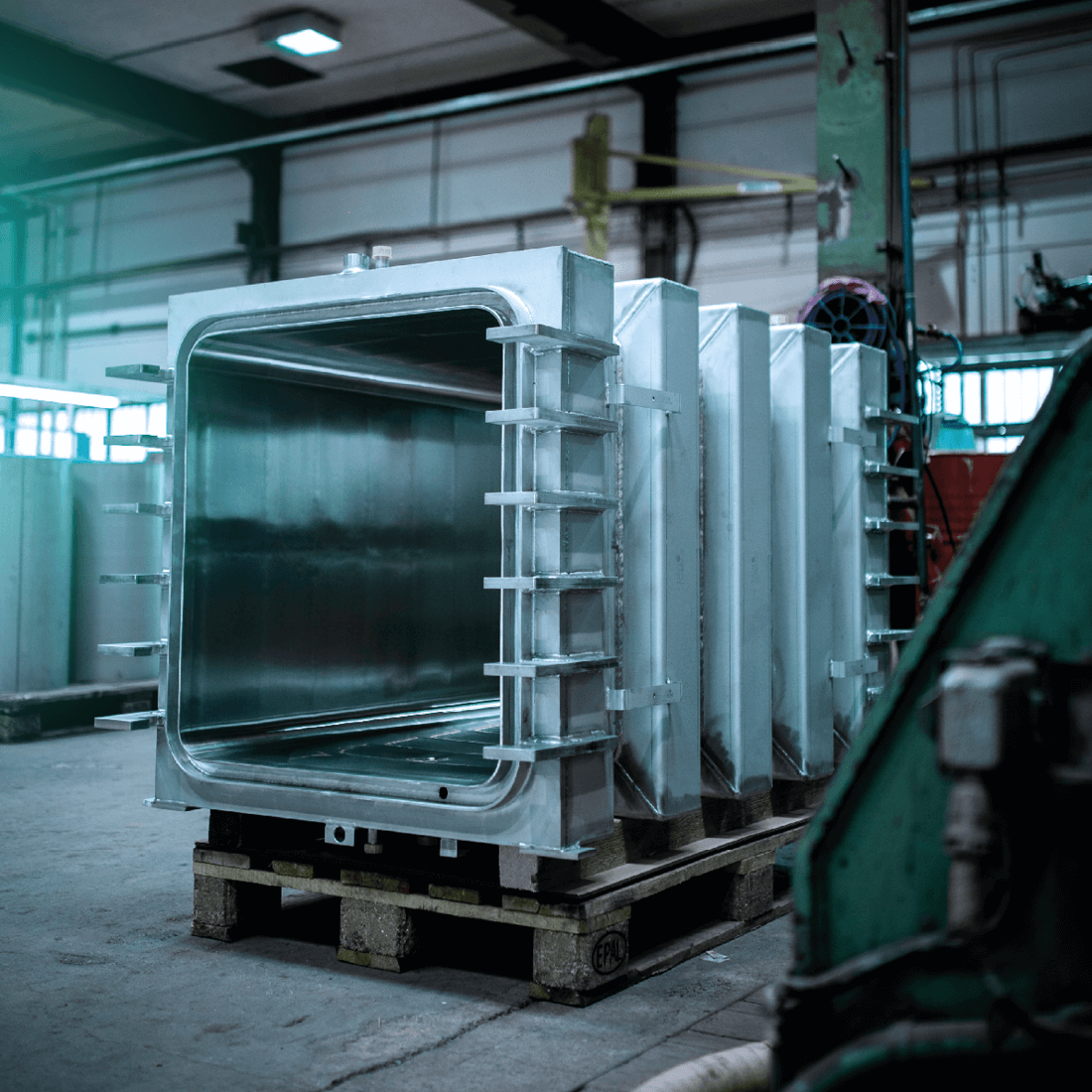 Several shiny stainless steel tanks with rectangular flanges on wooden pallets in an industrial hall.