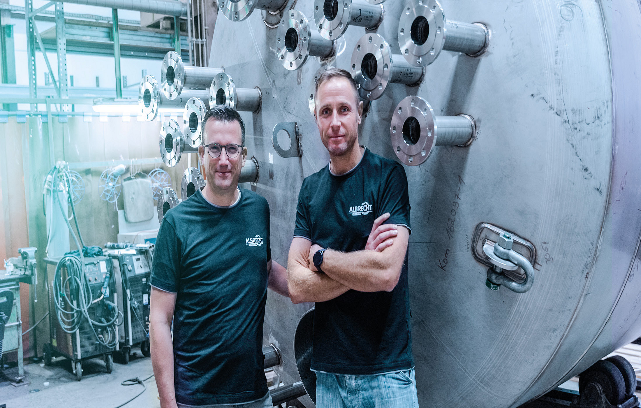 Zwei Mitarbeiter in Firmenkleidung posieren stolz vor einem großen Industriebehälter aus Metall.