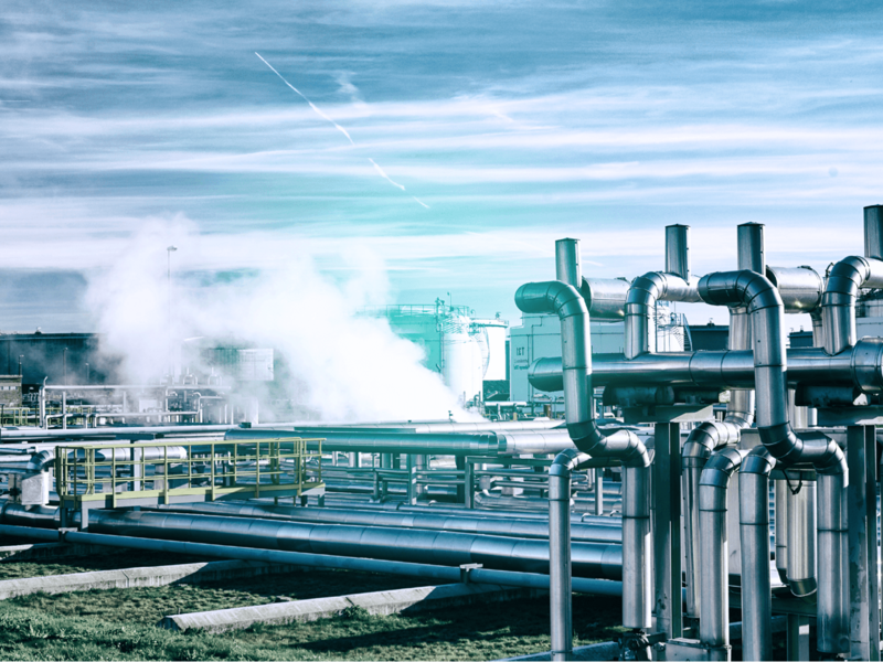 Komplexe Industrieanlage mit vielen Metallrohren und aufsteigendem Dampf unter blauem Himmel.