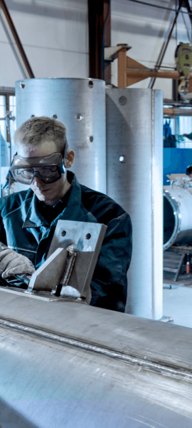 A craftsman uses a tool to grind welds on a large stainless steel pipe in a workshop.