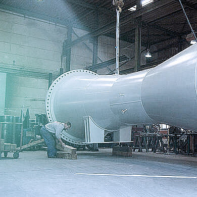 Arbeiter in einer Fabrik positionieren ein riesiges, schweres Metallrohr, das an einem Kran hängt.