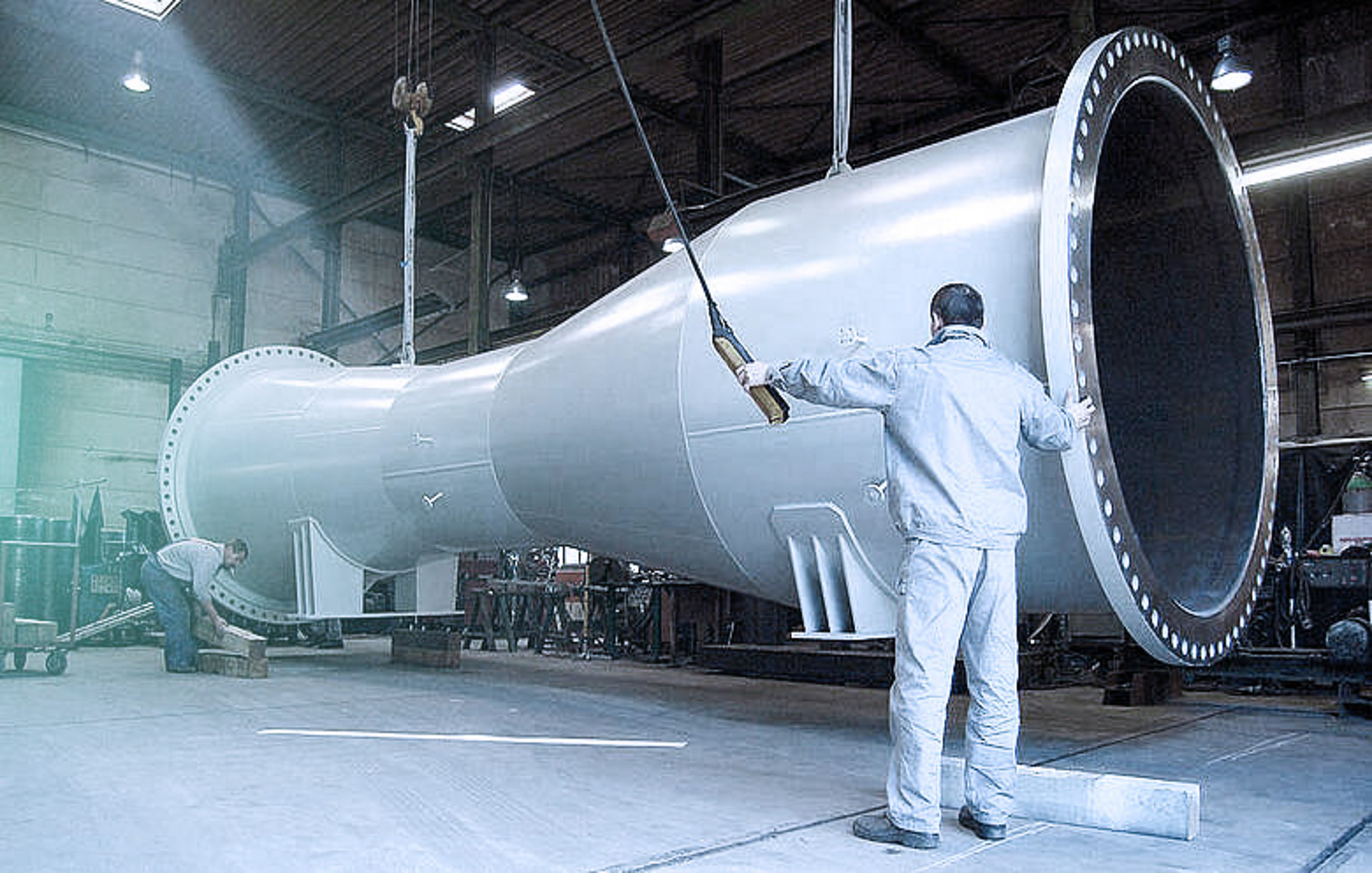 Arbeiter in einer Fabrik positionieren ein riesiges, schweres Metallrohr, das an einem Kran hängt.