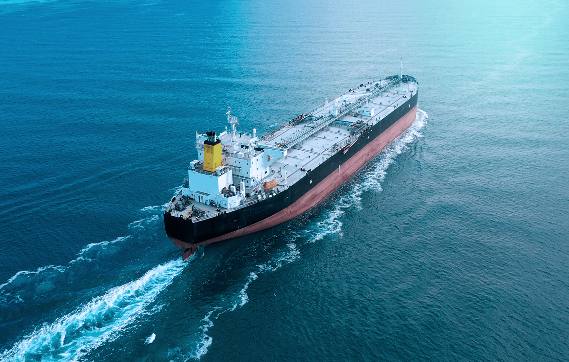 Aerial view of a large cargo ship sailing through the calm blue sea, leaving a white foam trail behind.
