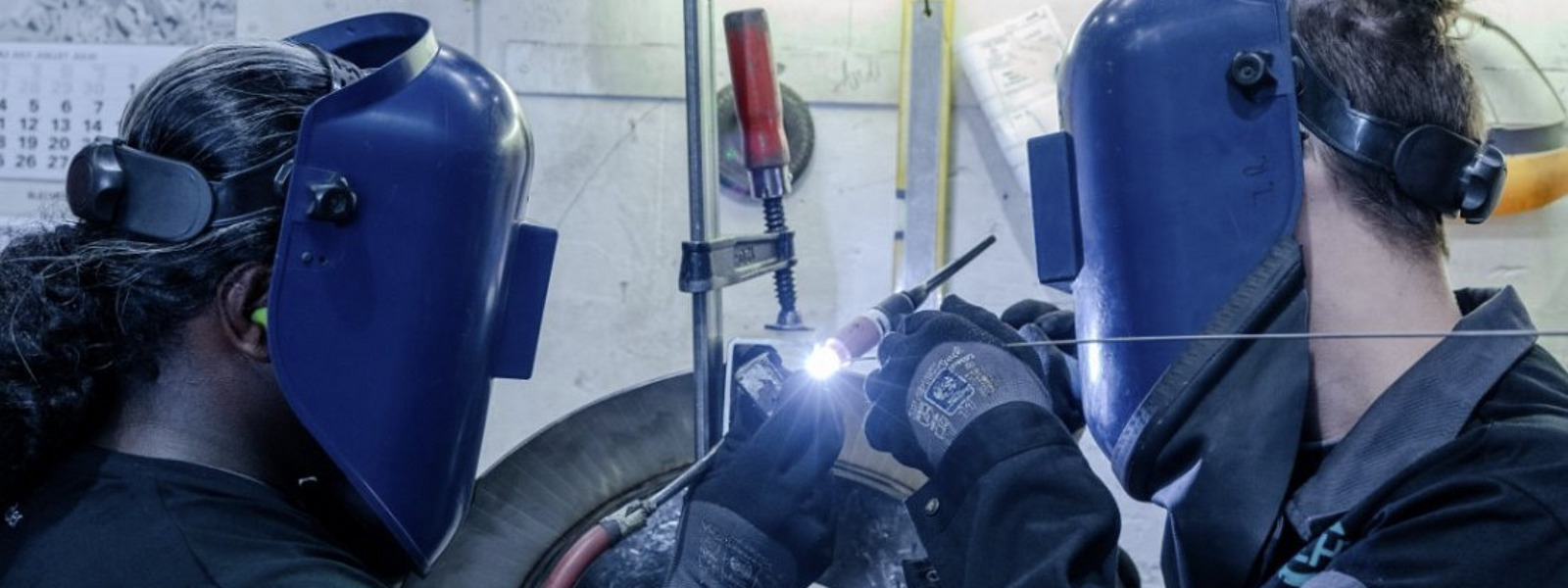 Zwei Personen in Schutzkleidung und mit Schweißermasken arbeiten konzentriert an einem Werkstück mit heller Schweißflamme.