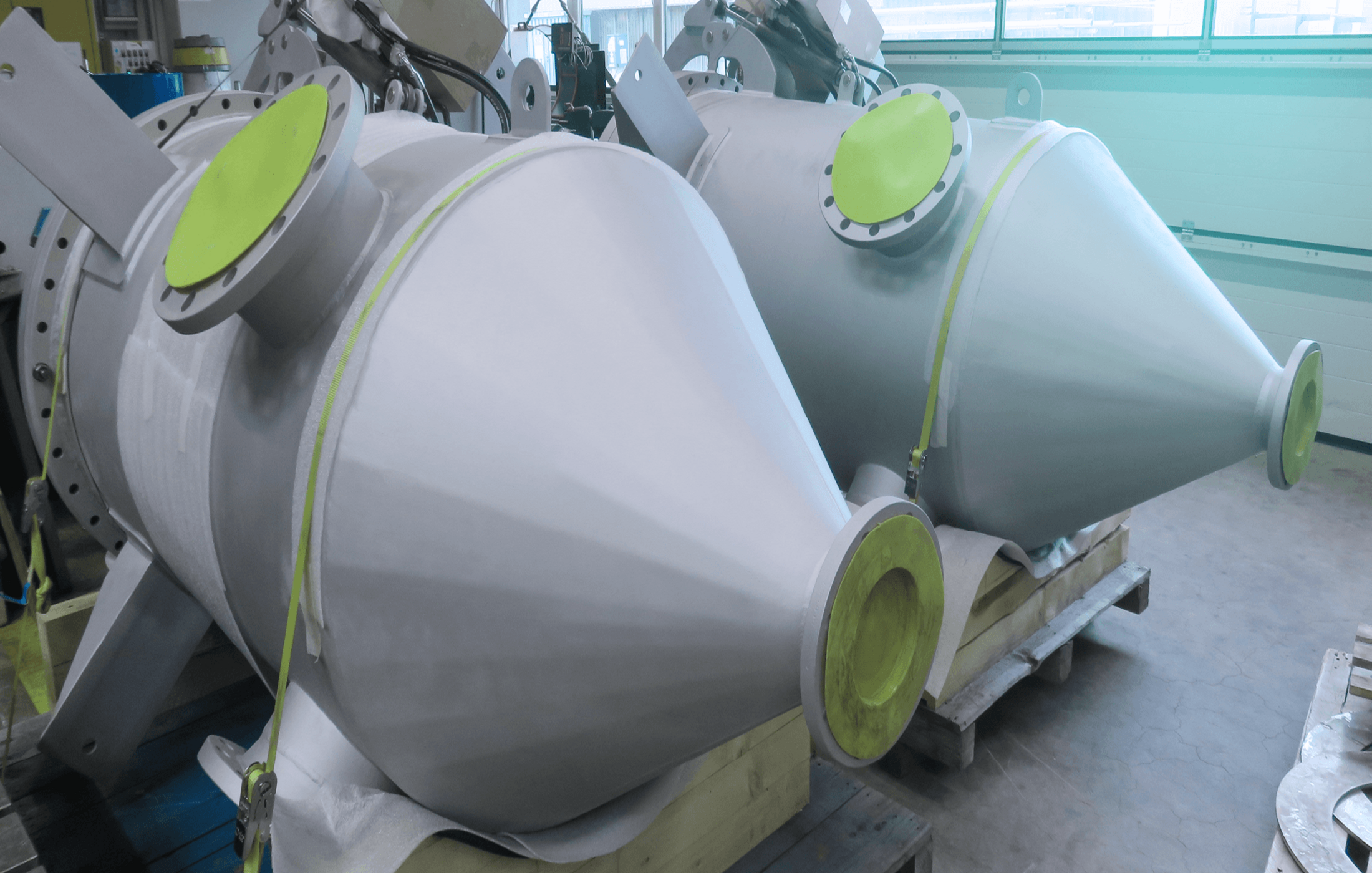 Two large conical stainless steel tanks with flanged openings on wooden pallets in a workshop.