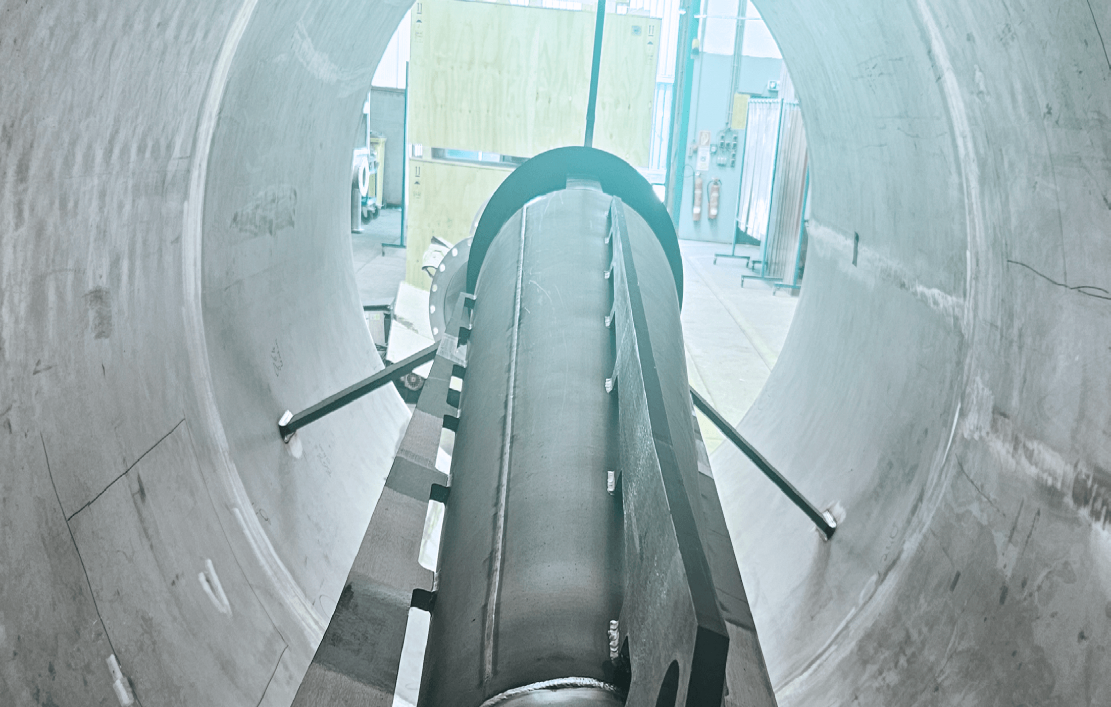 Blick durch ein großes, rundes Betonrohr auf eine zylindrische Stahlkonstruktion im industriellen Umfeld.