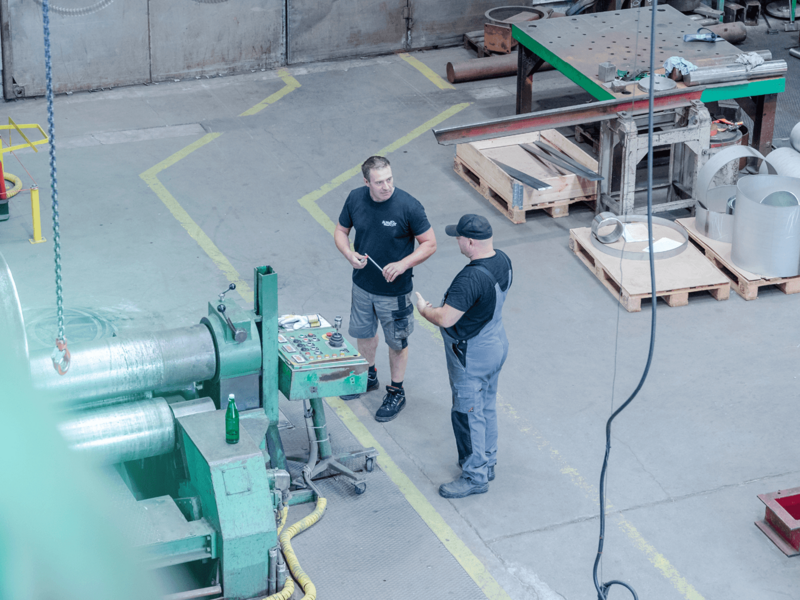 Two workers chat by an industrial machine in a large workshop hall with metal parts and workbenches.