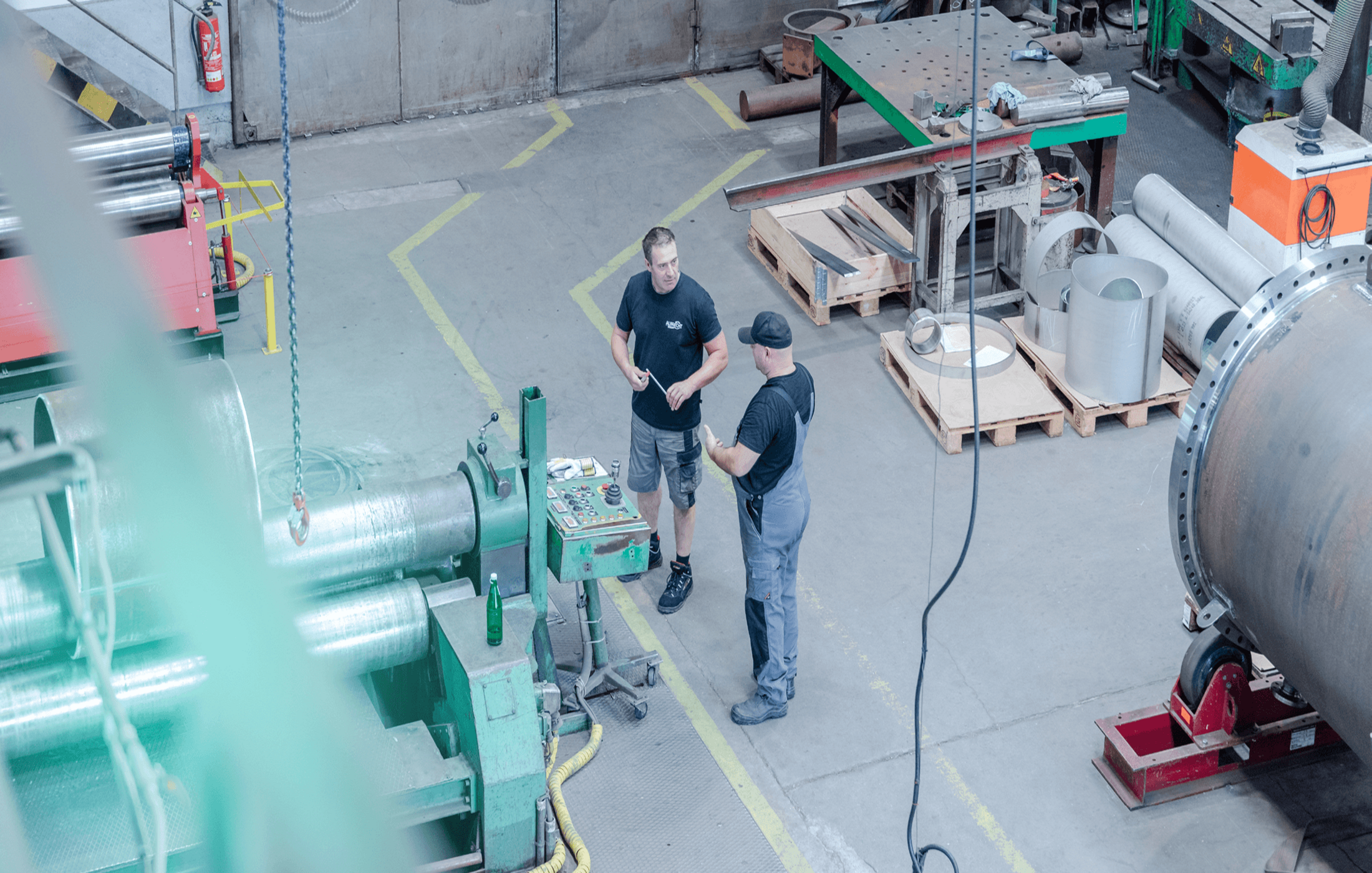 Zwei Arbeiter unterhalten sich an einer Industriemaschine in einer großen Werkhalle mit Metallteilen und Werkbänken.
